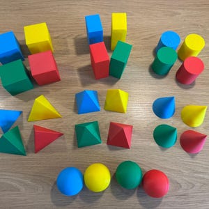 Peut inclure: Ensemble de formes géométriques colorées, comprenant des cubes, des cylindres, des cônes, des pyramides et des sphères. Les formes sont de différentes couleurs : bleu, jaune, vert et rouge. Les objets sont disposés sur une surface en bois.