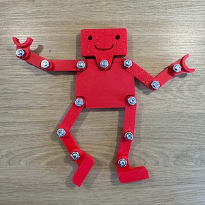 Peut inclure: Un jouet robot articulé rouge avec un visage souriant. Le robot a des bras et des jambes mobiles, reliés par des boulons en métal.