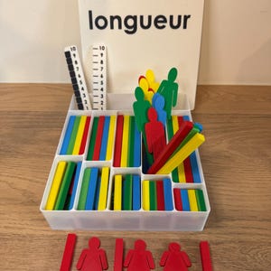 Op de afbeelding: Een witte plastic doos met vakken gevuld met gekleurde houten staafjes en uitgesneden figuren. De doos is gelabeld "Kit longueur".