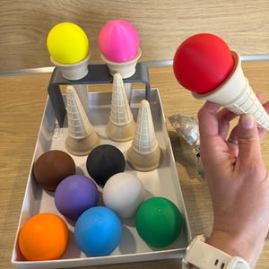 Peut inclure: Un ensemble de cônes de crème glacée jouets colorés. L'ensemble comprend des sphères de différentes couleurs, dont le jaune, le rose, le rouge, le marron, le noir, le violet, le blanc, le bleu et le vert. Les cônes sont beiges et l'ensemble est dans un plateau blanc.