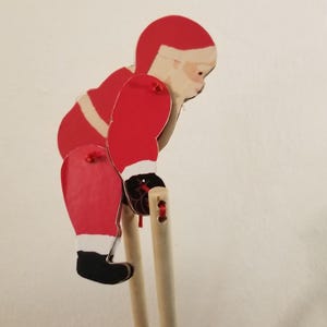 May include: A wooden Santa Claus figure, painted in red, white, and black, with a white beard and a red hat. The figure is bent over, with its arms extended, and is attached to two wooden dowels.