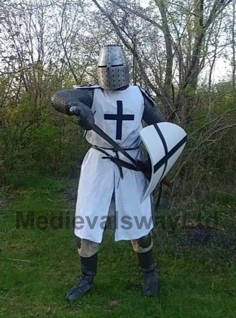 May include: A person in a medieval knight costume, including a metal helmet, chainmail, and a white tunic with a black cross. They are holding a spear and a shield with a black cross design. The background is a grassy area with trees.