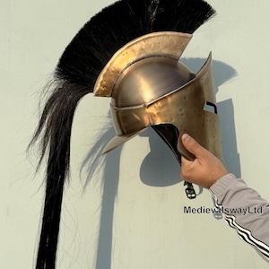 May include: A bronze Spartan helmet with a black horsehair plume. The helmet has a face mask and a raised crest. The helmet is held in a hand.