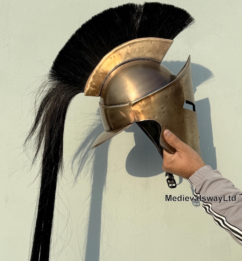 May include: A bronze Spartan helmet with a black horsehair plume. The helmet has a face mask and a raised crest. The helmet is held in a hand.