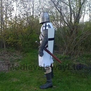 May include: A medieval knight in full armor stands in a grassy area. The knight wears a metal helmet, chainmail, and a white tunic with a black cross. A shield with a black cross and a sword are also visible.