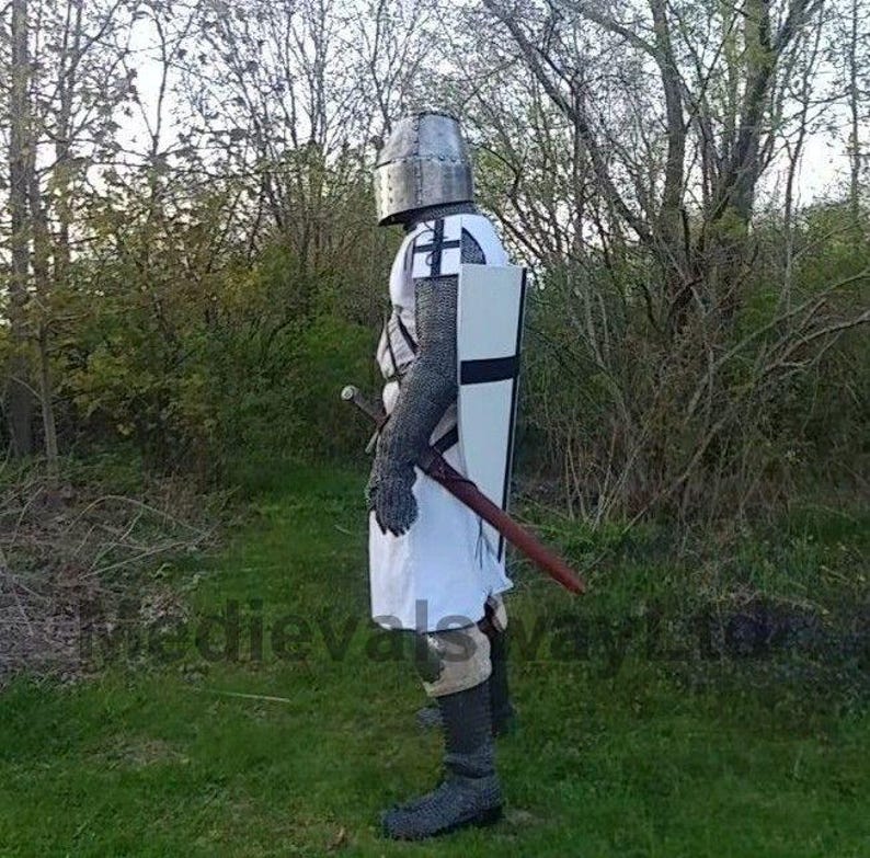 May include: A medieval knight in full armor stands in a grassy area. The knight wears a metal helmet, chainmail, and a white tunic with a black cross. A shield with a black cross and a sword are also visible.