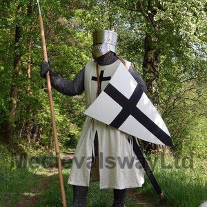 May include: A medieval knight in full armor stands in a forest, holding a spear. The knight wears a helmet, chainmail, and a white tunic with a black cross. A white shield with a black cross is held in the other hand.