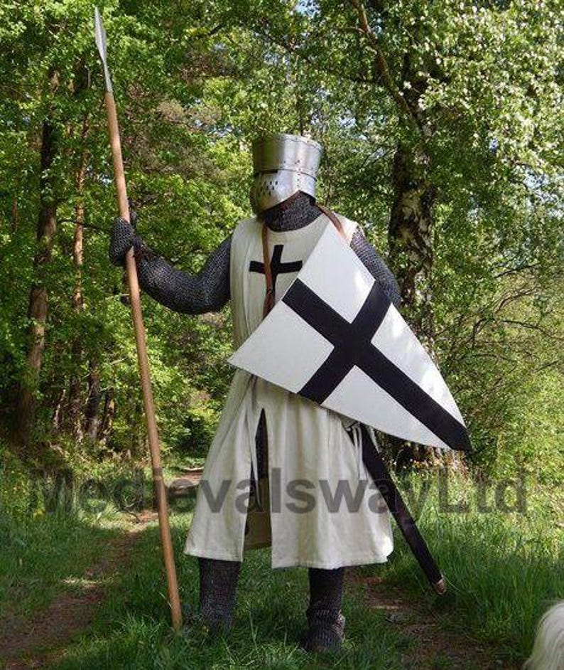 May include: A medieval knight in full armor stands in a forest, holding a spear. The knight wears a helmet, chainmail, and a white tunic with a black cross. A white shield with a black cross is held in the other hand.