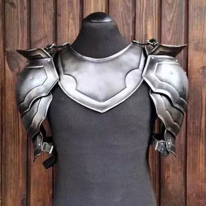 Könnte beinhalten: Ein Paar silberfarbene Schulterpanzer aus Metall mit einem dekorativen Design. Die Rüstung wird von einer schwarzen Schaufensterpuppe getragen.
