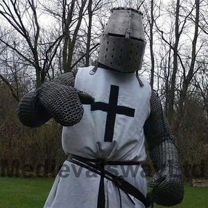 May include: A medieval knight's costume featuring a metal helmet, chainmail, and a white tunic with a black cross. The costume includes chainmail gloves and a black belt. The background shows bare trees.