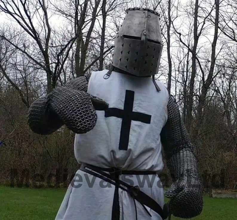 May include: A medieval knight's costume featuring a metal helmet, chainmail, and a white tunic with a black cross. The costume includes chainmail gloves and a black belt. The background shows bare trees.