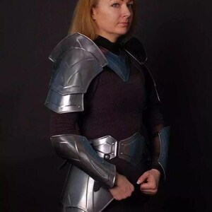 May include: A woman wearing silver armor with a black shirt and pants. The armor includes shoulder pads, bracers, and a belt.