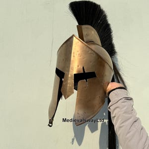 May include: A gold-colored Spartan helmet with a black horsehair plume. The helmet has a distinctive design with a raised crest and a face mask with a narrow eye slit.