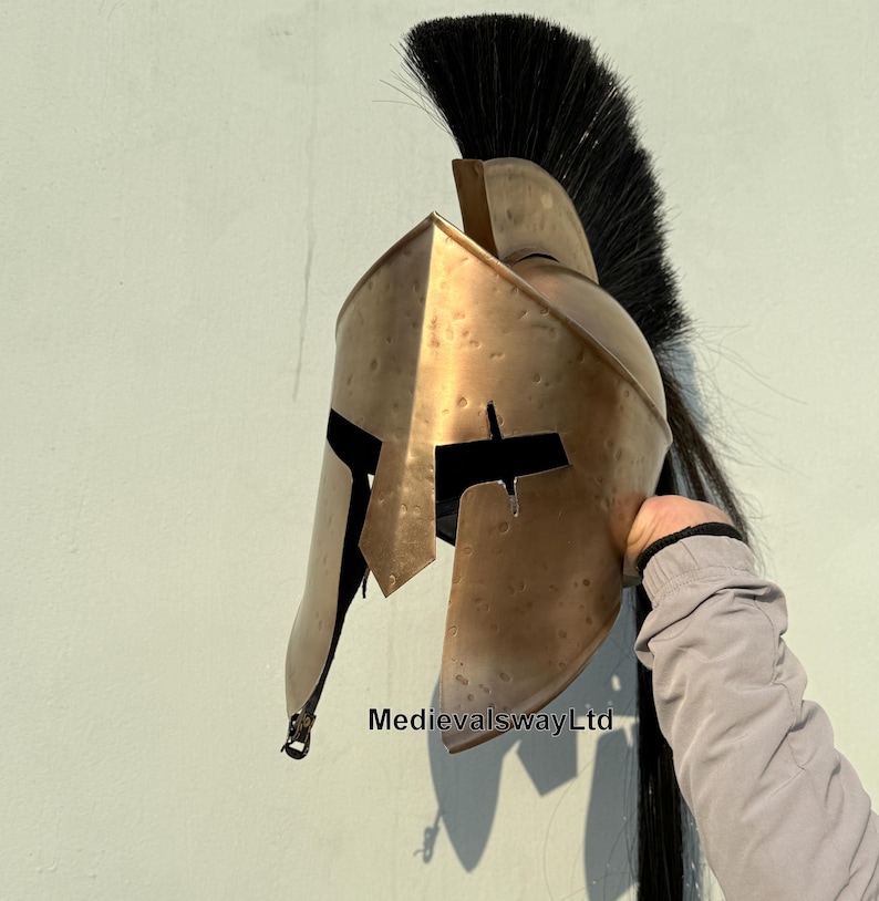 May include: A gold-colored Spartan helmet with a black horsehair plume. The helmet has a distinctive design with a raised crest and a face mask with a narrow eye slit.