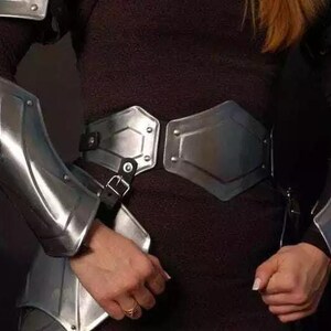 May include: A silver metal armor set with a chest plate, arm guards, and a belt. The armor is worn by a person with long, blonde hair.