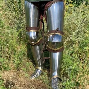 May include: Polished steel leg armor with leather straps and brass accents. The armor is highly reflective, with a bright, metallic sheen. The knee and ankle areas are articulated for movement. The armor is designed for protection and historical reenactment.