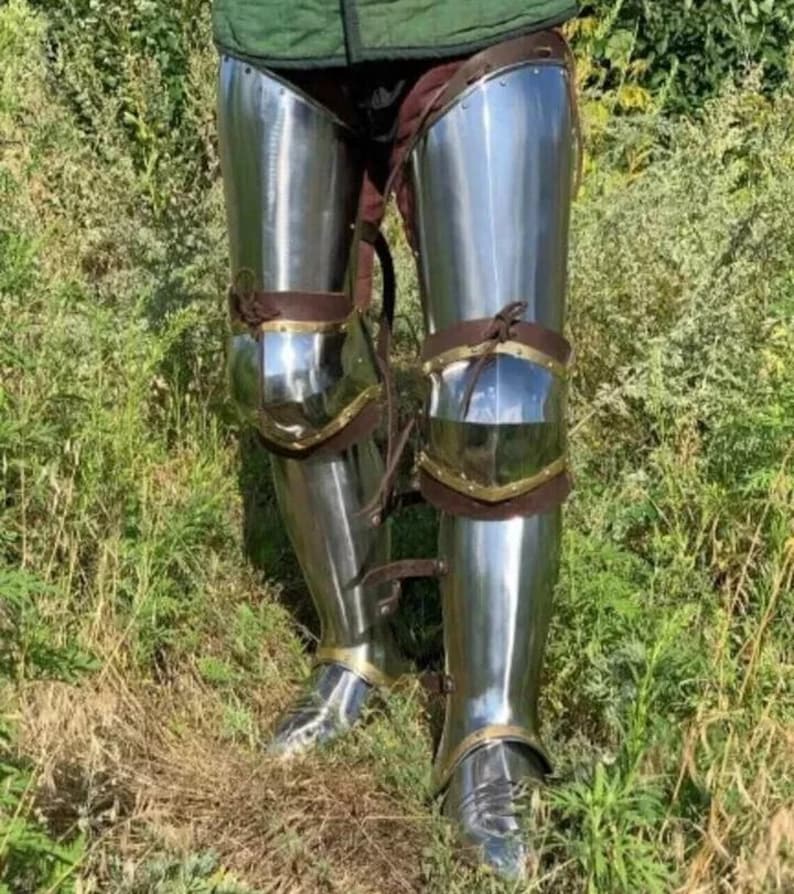 May include: Polished steel leg armor with leather straps and brass accents. The armor is highly reflective, with a bright, metallic sheen. The knee and ankle areas are articulated for movement. The armor is designed for protection and historical reenactment.