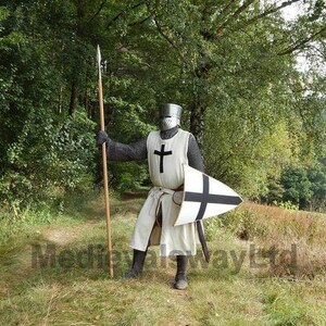 May include: A medieval knight in full armor stands in a grassy area, holding a spear and shield. The knight wears a white tunic with a black cross, chainmail, and a helmet. The shield is white with a black cross. The background features trees.