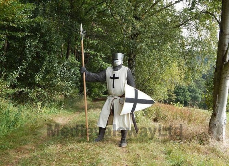 May include: A medieval knight in full armor stands in a grassy area, holding a spear and shield. The knight wears a white tunic with a black cross, chainmail, and a helmet. The shield is white with a black cross. The background features trees.
