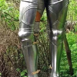 Puede incluir: Un par de armaduras de pierna de metal plateado con correas y hebillas. La armadura se lleva sobre los pantalones y cubre las piernas desde el muslo hasta el pie.