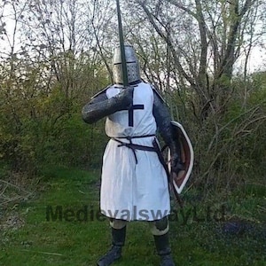 May include: A medieval knight in full armor, including a helmet, chainmail, and white tunic with a black cross. The knight holds a sword and shield, standing in a grassy area with trees in the background. The image has the text "MedievalswayLtd".