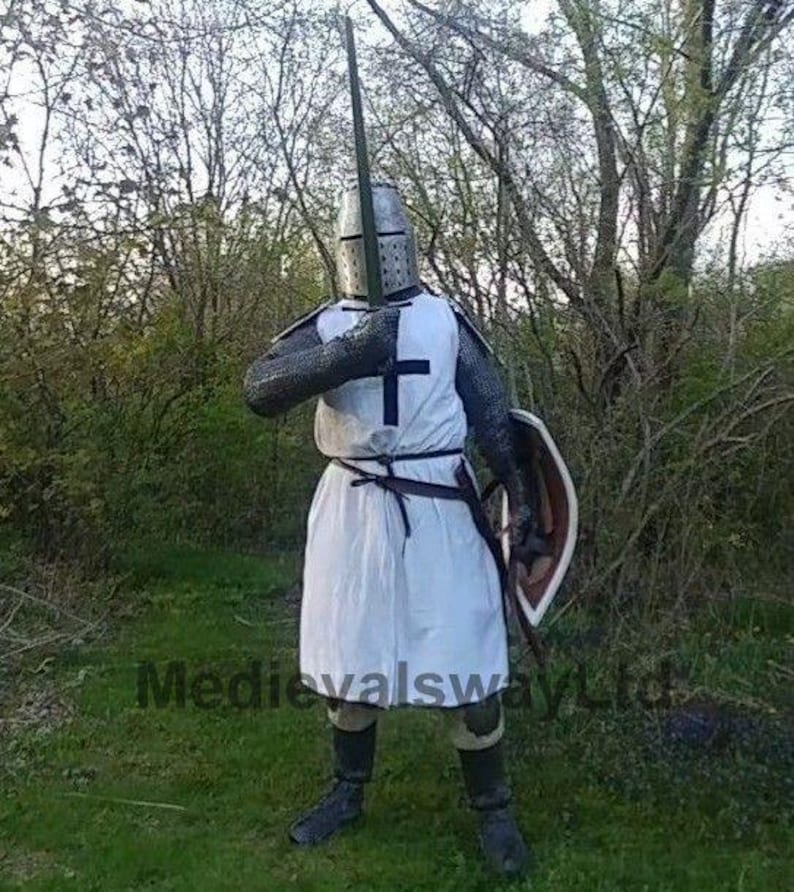 May include: A medieval knight in full armor, including a helmet, chainmail, and white tunic with a black cross. The knight holds a sword and shield, standing in a grassy area with trees in the background. The image has the text "MedievalswayLtd".