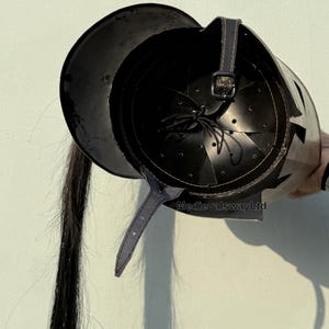 May include: Black leather helmet with a long black hair tail. The helmet has a pointed crown and a decorative design on the side. The helmet is made of leather and has a strap to secure it to the head. MedievalswayLtd is printed on the helmet.