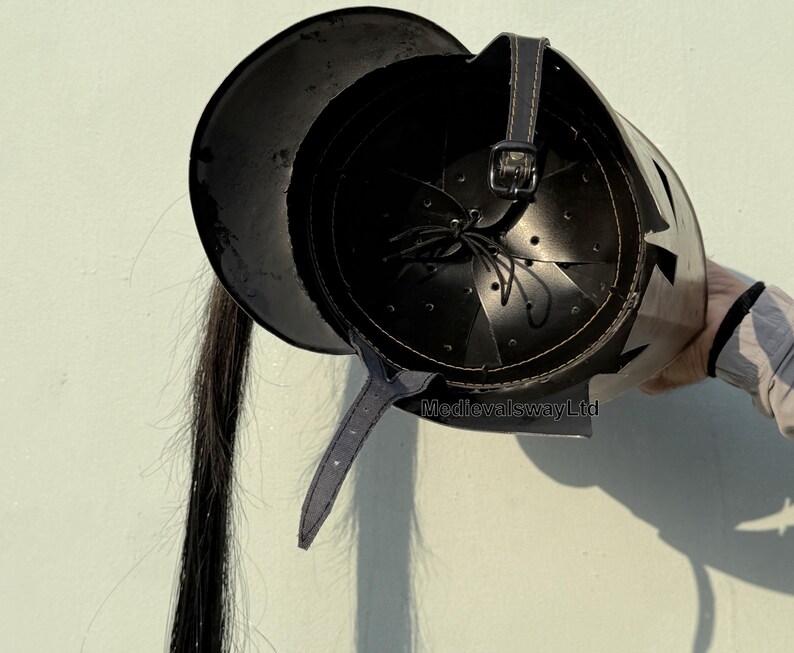 May include: Black leather helmet with a long black hair tail. The helmet has a pointed crown and a decorative design on the side. The helmet is made of leather and has a strap to secure it to the head. MedievalswayLtd is printed on the helmet.