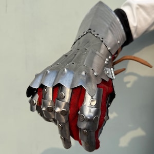 K&ouml;nnte beinhalten: Ein mittelalterlicher Panzerhandschuh aus polierten Stahlplatten, die vernietet sind. Der Handschuh hat bewegliche Fingerplatten und eine ausgestellte Manschette. Die Innenseite ist mit rotem Stoff gef&uuml;ttert. Der Handschuh dient zum Schutz von Hand und Handgelenk.