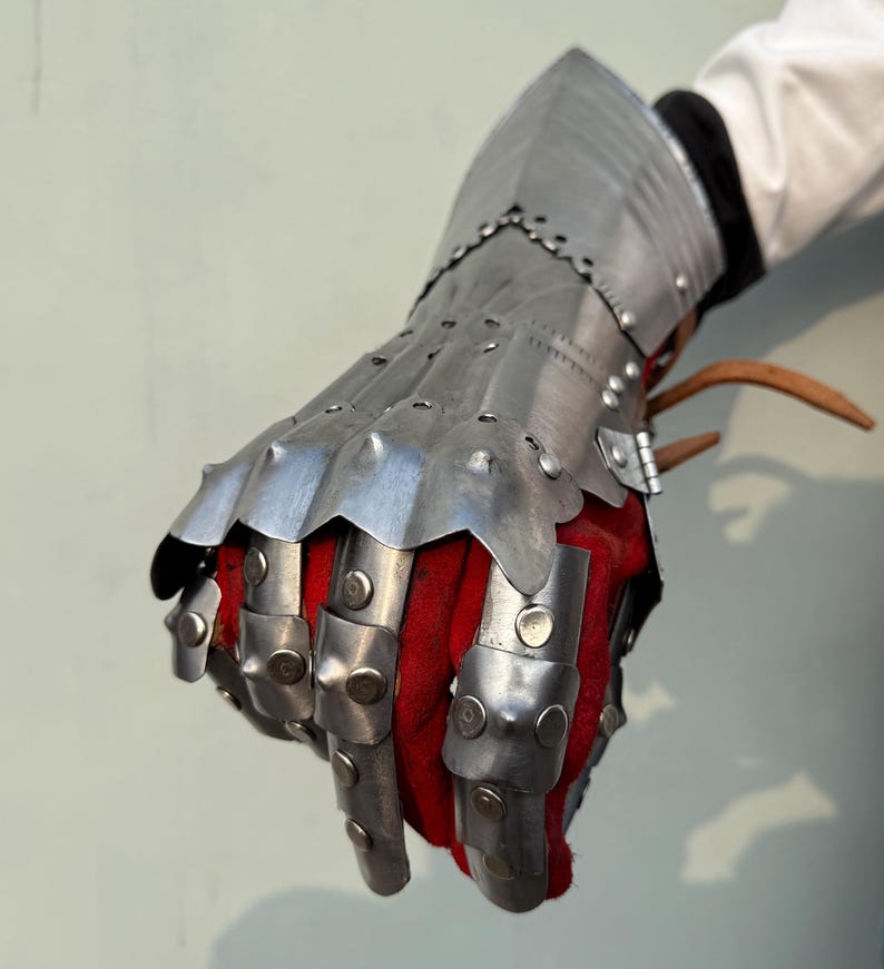K&ouml;nnte beinhalten: Ein mittelalterlicher Panzerhandschuh aus polierten Stahlplatten, die vernietet sind. Der Handschuh hat bewegliche Fingerplatten und eine ausgestellte Manschette. Die Innenseite ist mit rotem Stoff gef&uuml;ttert. Der Handschuh dient zum Schutz von Hand und Handgelenk.
