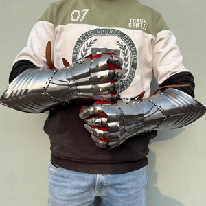 K&ouml;nnte beinhalten: Ein Paar polierte Stahlhandschuhe mit beweglichen Fingern und rotem Stofffutter. Die Handschuhe sind zum Schutz der H&auml;nde und Unterarme konzipiert. Die Metallr&uuml;stung ist vernietet und das Design erinnert an mittelalterliche R&uuml;stungen.