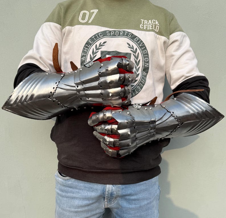 K&ouml;nnte beinhalten: Ein Paar polierte Stahlhandschuhe mit beweglichen Fingern und rotem Stofffutter. Die Handschuhe sind zum Schutz der H&auml;nde und Unterarme konzipiert. Die Metallr&uuml;stung ist vernietet und das Design erinnert an mittelalterliche R&uuml;stungen.