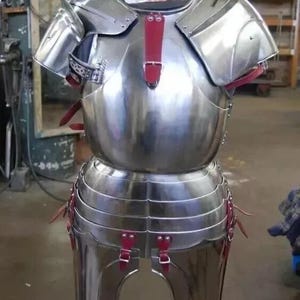 May include: A silver metal suit of armour with red leather straps. The armour is made up of a breastplate, shoulder pads, and a skirt. The armour is displayed on a wooden stand.