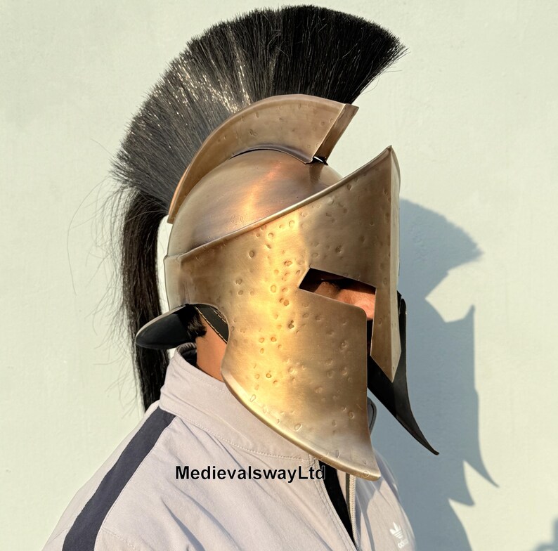 May include: A gold-colored Spartan helmet with a black horsehair plume. The helmet has a distinctive curved shape and a face mask with a narrow eye slit.
