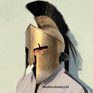 May include: A gold-colored Spartan helmet with a black horsehair plume. The helmet has a distinctive design with a raised crest and a face mask with a narrow opening for the eyes. The helmet is worn by a person, but the person's face is not visible.