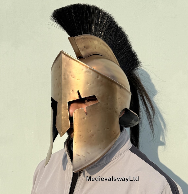 May include: A gold-colored Spartan helmet with a black horsehair plume. The helmet has a distinctive design with a raised crest and a face mask with a narrow opening for the eyes. The helmet is worn by a person, but the person's face is not visible.