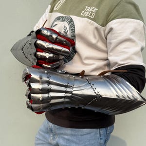 K&ouml;nnte beinhalten: Ein Paar polierte Stahlhandschuhe mit beweglichen Fingern und roter Innenseite. Die Handschuhe dienen zum Schutz der H&auml;nde und Handgelenke. Das Bild zeigt die Handschuhe vor hellem Hintergrund.