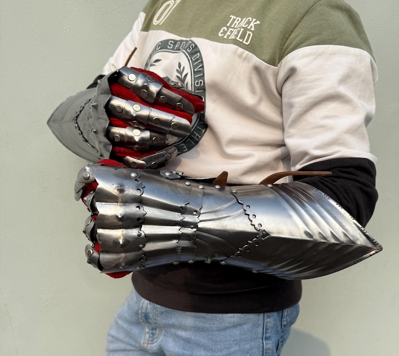 K&ouml;nnte beinhalten: Ein Paar polierte Stahlhandschuhe mit beweglichen Fingern und roter Innenseite. Die Handschuhe dienen zum Schutz der H&auml;nde und Handgelenke. Das Bild zeigt die Handschuhe vor hellem Hintergrund.