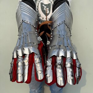 K&ouml;nnte beinhalten: Ein Paar Stahlhandschuhe mit beweglichen Fingern und roter Innenseite. Die Handschuhe bestehen aus genieteten Metallplatten und sollen die H&auml;nde sch&uuml;tzen. Das Design erinnert an mittelalterliche R&uuml;stungen.
