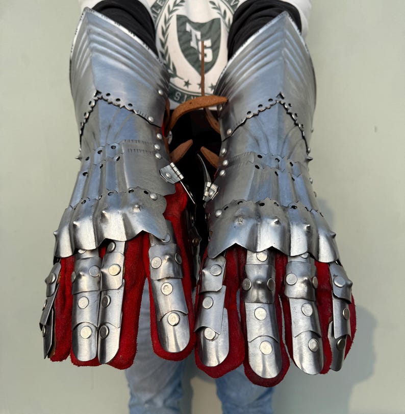 K&ouml;nnte beinhalten: Ein Paar Stahlhandschuhe mit beweglichen Fingern und roter Innenseite. Die Handschuhe bestehen aus genieteten Metallplatten und sollen die H&auml;nde sch&uuml;tzen. Das Design erinnert an mittelalterliche R&uuml;stungen.