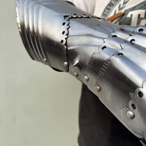 K&ouml;nnte beinhalten: Nahaufnahme eines mittelalterlichen Stahlhandschuhs, der sein gegliedertes Design zeigt. Der Handschuh besteht aus poliertem Metall mit Nieten und dekorativen Kanten. Die R&uuml;stung scheint gut verarbeitet zu sein, Licht zu reflektieren und Haltbarkeit zu suggerieren.