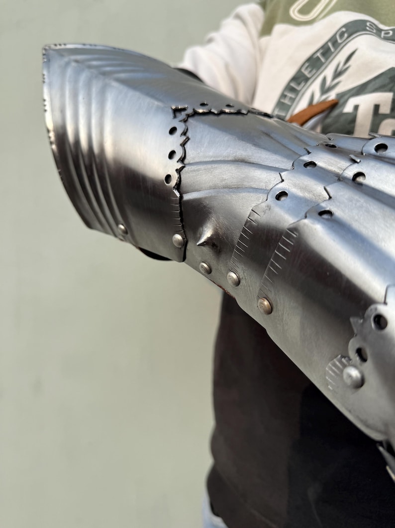 K&ouml;nnte beinhalten: Nahaufnahme eines mittelalterlichen Stahlhandschuhs, der sein gegliedertes Design zeigt. Der Handschuh besteht aus poliertem Metall mit Nieten und dekorativen Kanten. Die R&uuml;stung scheint gut verarbeitet zu sein, Licht zu reflektieren und Haltbarkeit zu suggerieren.
