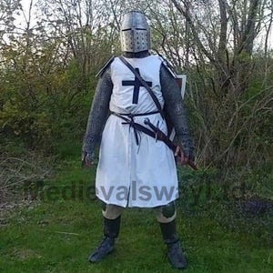 May include: A full-body shot of a person in medieval armor. The outfit includes a metal helmet, chainmail, a white tunic with a black cross, and a sword. The person is standing in a grassy area with trees in the background.