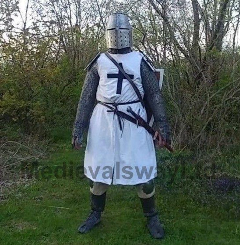 May include: A full-body shot of a person in medieval armor. The outfit includes a metal helmet, chainmail, a white tunic with a black cross, and a sword. The person is standing in a grassy area with trees in the background.