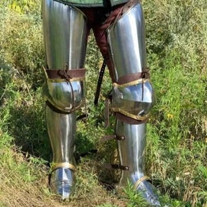 May include: Polished steel leg armor, reflecting the surrounding green grass and foliage. The armor includes knee protection and is secured with brown leather straps and brass accents. The medieval-style armor is designed for protection and display.