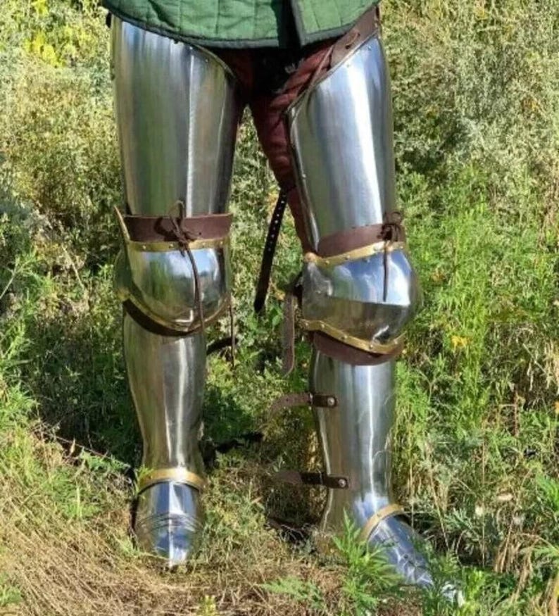 May include: Polished steel leg armor, reflecting the surrounding green grass and foliage. The armor includes knee protection and is secured with brown leather straps and brass accents. The medieval-style armor is designed for protection and display.