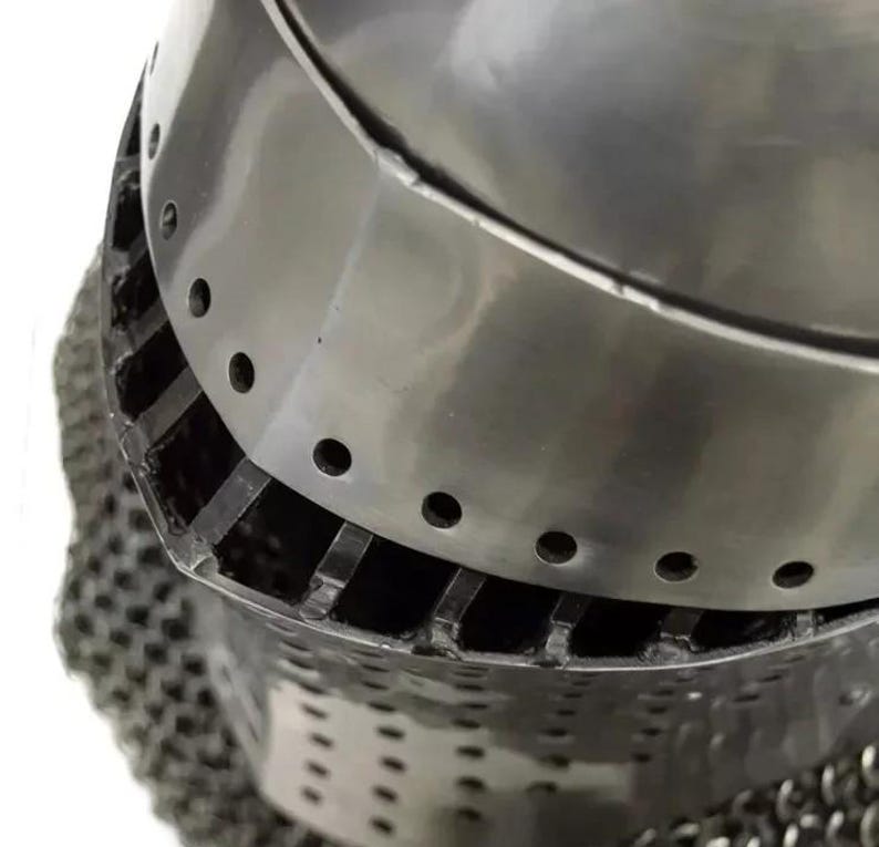 May include: Close-up of a medieval steel helmet with a polished finish. The helmet features a rounded top with a series of small, evenly spaced holes and a lower section with vertical slits and chainmail.