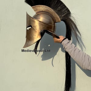 May include: A gold-colored Spartan helmet with a black horsehair plume. The helmet has a distinctive crest and a face mask with a small eye opening. The helmet is made of metal and has a distressed finish.