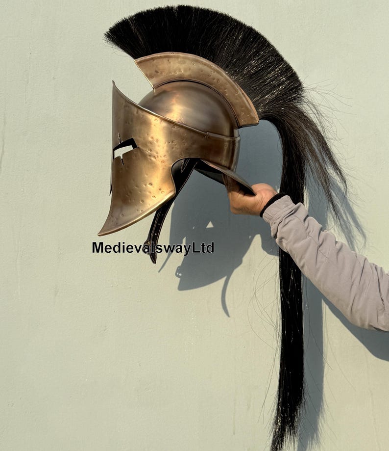 May include: A gold-colored Spartan helmet with a black horsehair plume. The helmet has a distinctive crest and a face mask with a small eye opening. The helmet is made of metal and has a distressed finish.