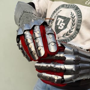 K&ouml;nnte beinhalten: Ein Paar gegliederte Stahlhandschuhe mit rotem Stofffutter. Die Handschuhe bestehen aus genieteten Metallplatten und dienen zum Schutz der H&auml;nde und Unterarme. Das Metall hat eine polierte, reflektierende Oberfl&auml;che.